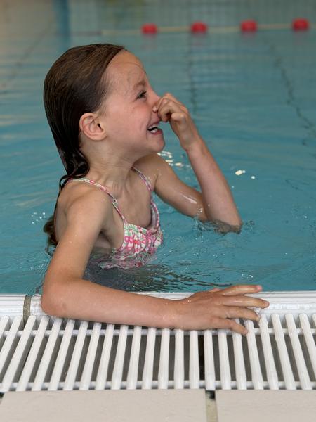 A young girl with wet hair is captured mid-action in a bright blue swimming pool, holding her nose with a playful grimace